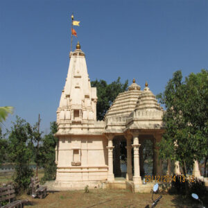 A traditional Hindu temple built from light-colored stone, featuring a tall shikhara spire with a yellow flag, an open pillared mandapa, and surrounding green trees under a bright blue sky.