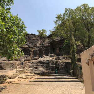 A view from the stone pathway looking up at the ancient rock-cut Pandava caves built into a rocky hillside with stairs and trees under a clear sky.