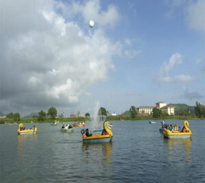 Several colorful swan and dragon-themed pedal boats carrying tourists across a wide lake with a fountain and a hilly background under a cloudy sky.