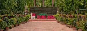 A wide panoramic view of a paved brick walkway lined with numerous potted plants and trees, leading to a large sign that reads "I AM IN WAGHAI BOTANICAL GARDEN" against a green living wall.